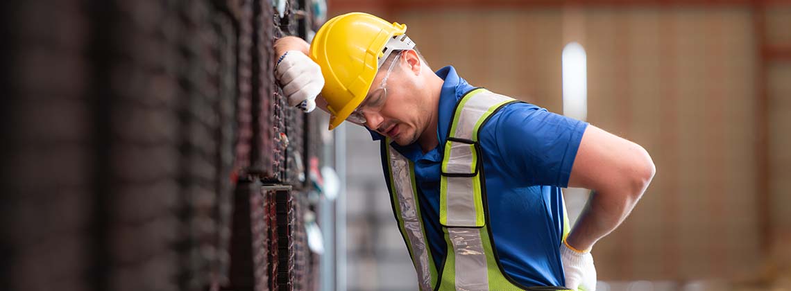 Male worker wearing a safety vest and helmet standing on a steels pallet due to back pain from working in a factory lifting heavy things.